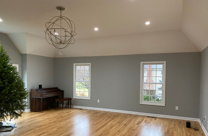 Spacious room with hardwood floors, a chandelier, and windows providing natural light, featuring a piano and a partially visible christmas tree.