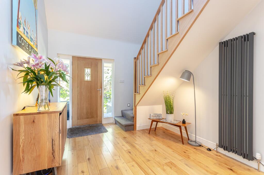 spacious hallway with oak staircase and flooring. oak sideboard with plants and flowers on it. oak front door with two side glass panels and a tall radiator adjacent to it