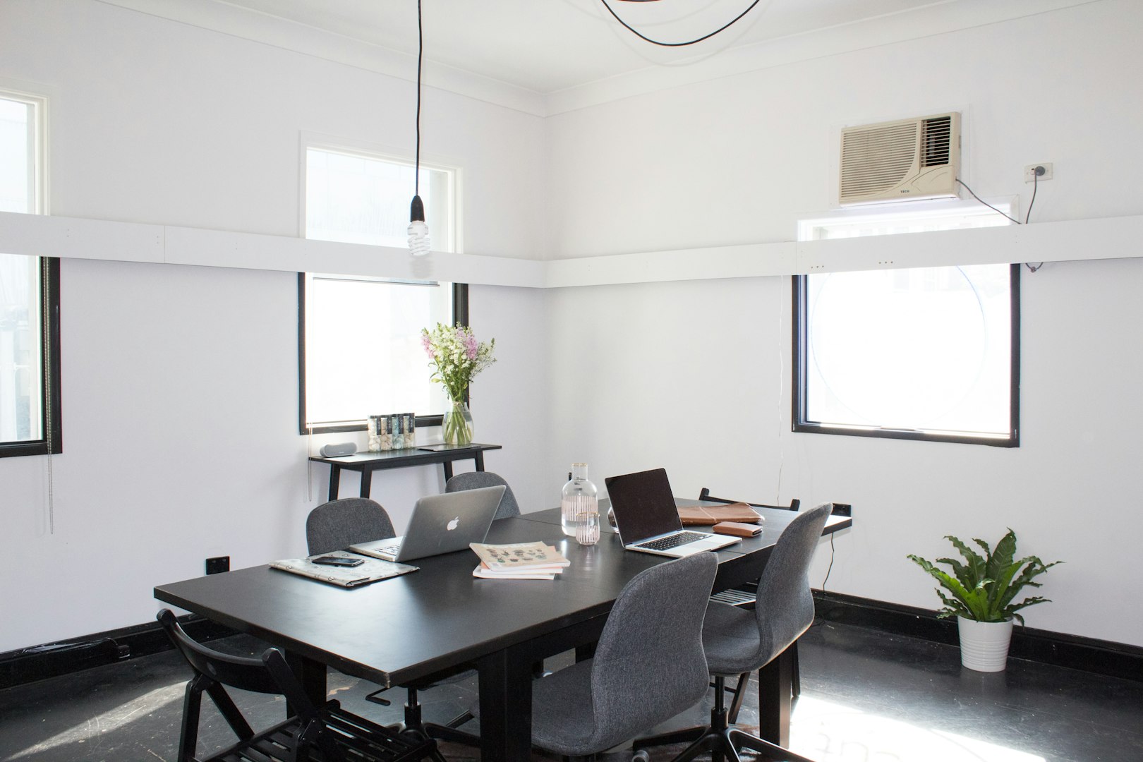 white modern office with a black desk and laptops sat on it. hanging from the ceiling there is a black lighting pendant with a large bulb. in the background there are windows and a small table with flowers on.